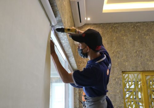 A worker using a drill to install window blinds in an indoor setting.